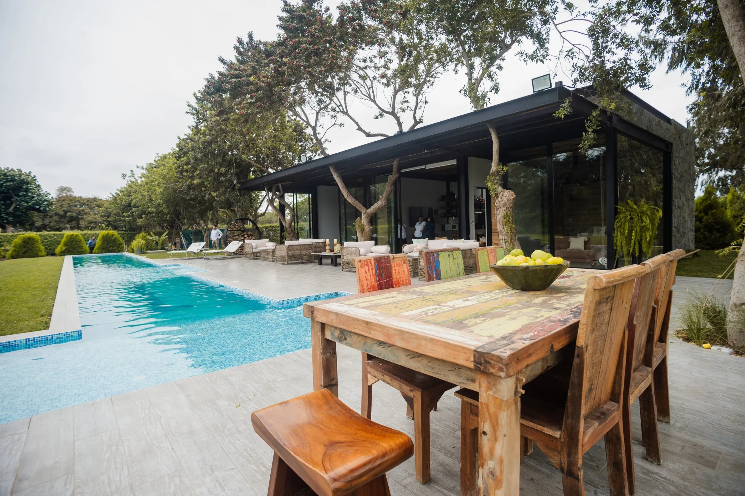 Comedor exterior de madera junto a la piscina, con la casa de vidrio al fondo.