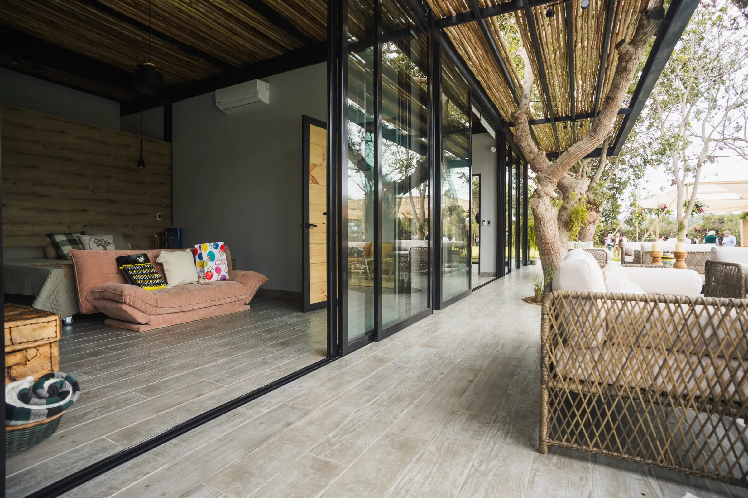 La fachada de vidrio abierta a la terraza, con el árbol, la pérgola y la sala de estar adentro.