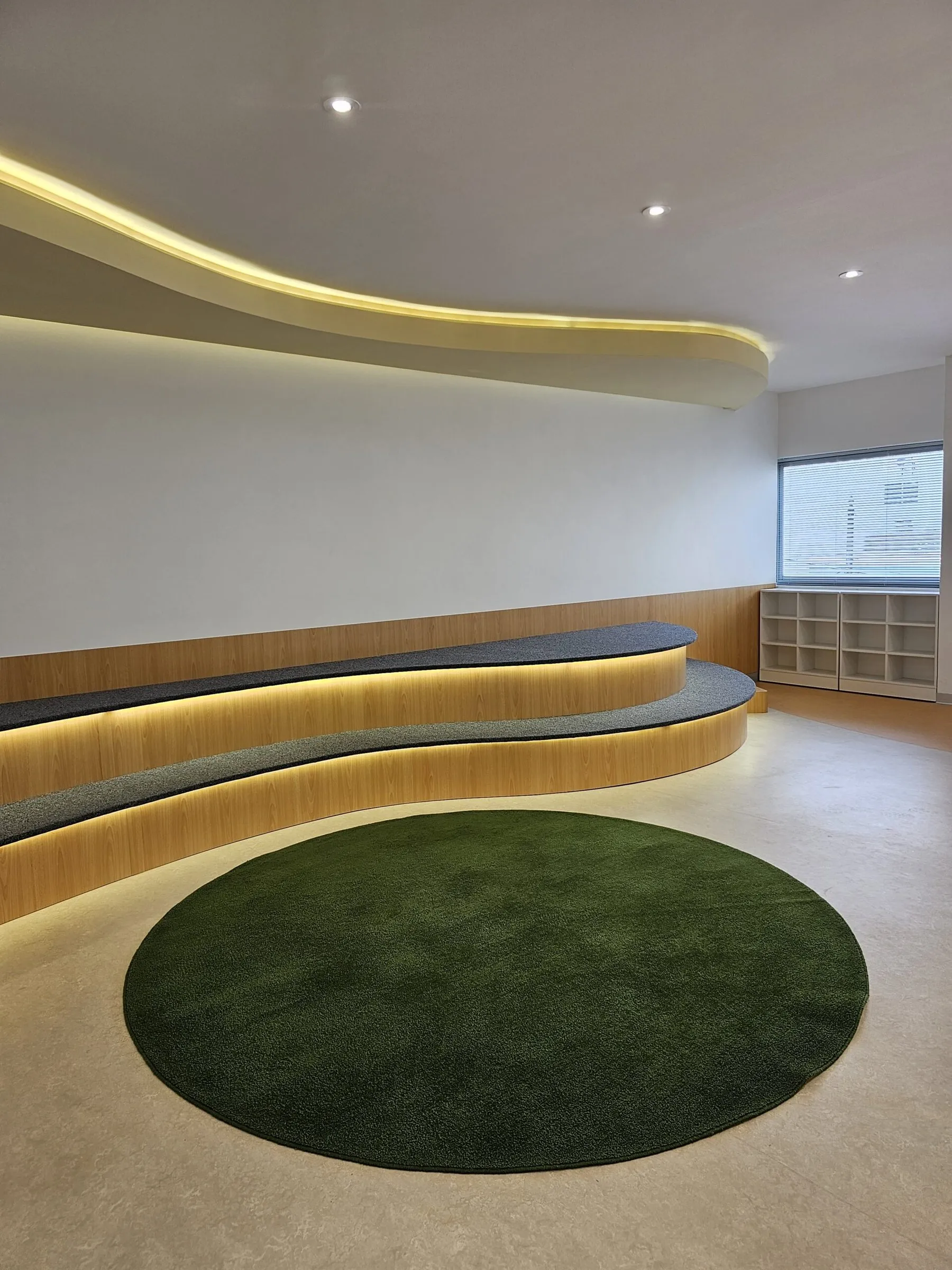 La gradería de madera con luz bajo el cielo raso ondulado, y una alfombra verde redonda.