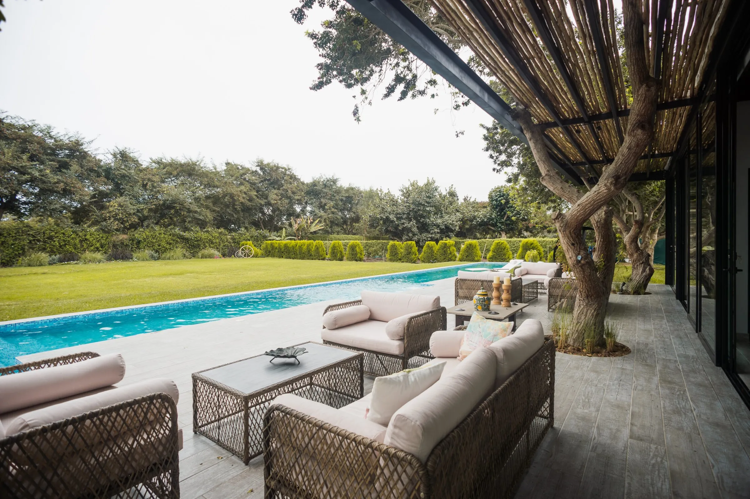 Sala exterior de mimbre bajo la pérgola, frente a la piscina y el jardín.